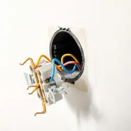 Rozpoczęta wymiana puszki elektrycznej. Kolorowe przewody wychodzą z czarnej obudowy w ścianie.