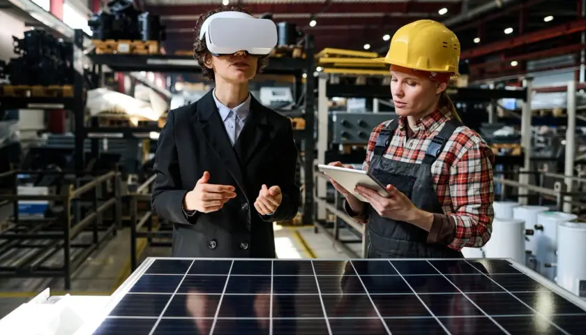 Kobieta w goglach VR i pracownica w kasku analizują panel słoneczny w fabryce.